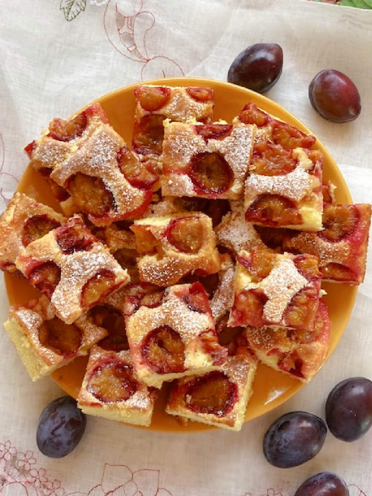 Overhead shot of a yellow plate with squares of plum cake on, and fresh plums around the plate.