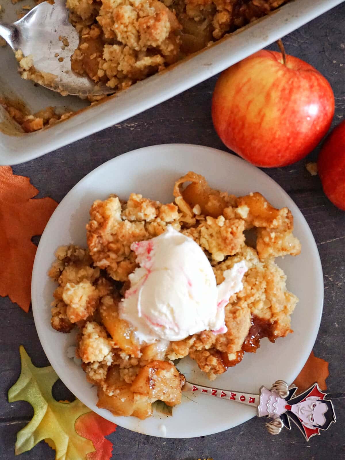 Overhead shoot of a white plate with apple crumble topped with a scoop of ice cream, 2 apples and a fish with more crumble at the top of the plate.
