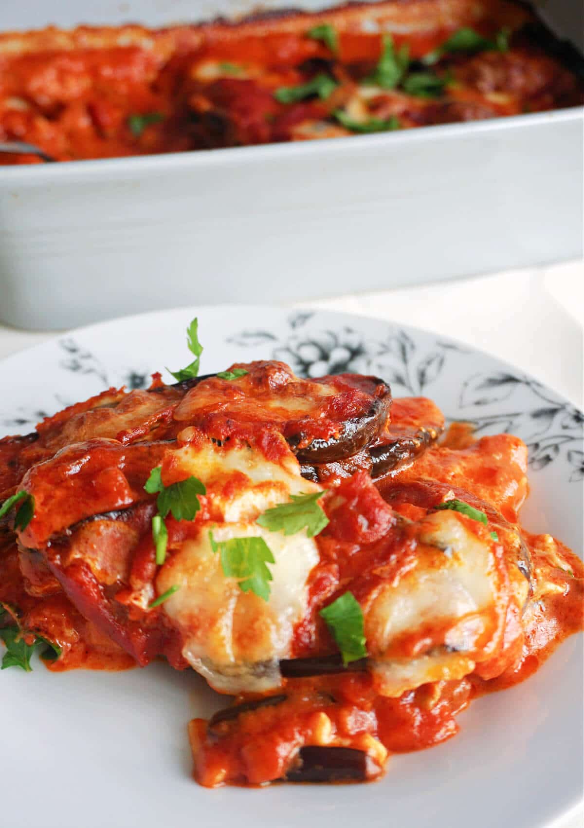 A white plate with a portion of eggplant parmesan and a dish in the background.