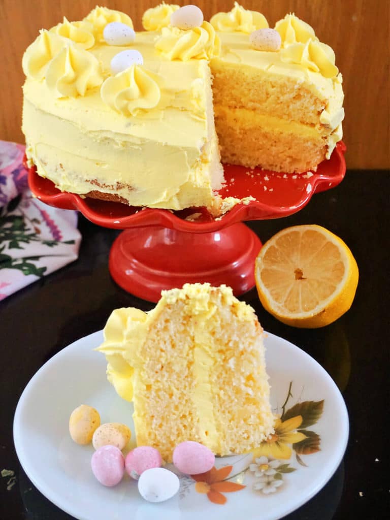 A slice of lemon cake on a white plate with more cake on a red cake stand in the background