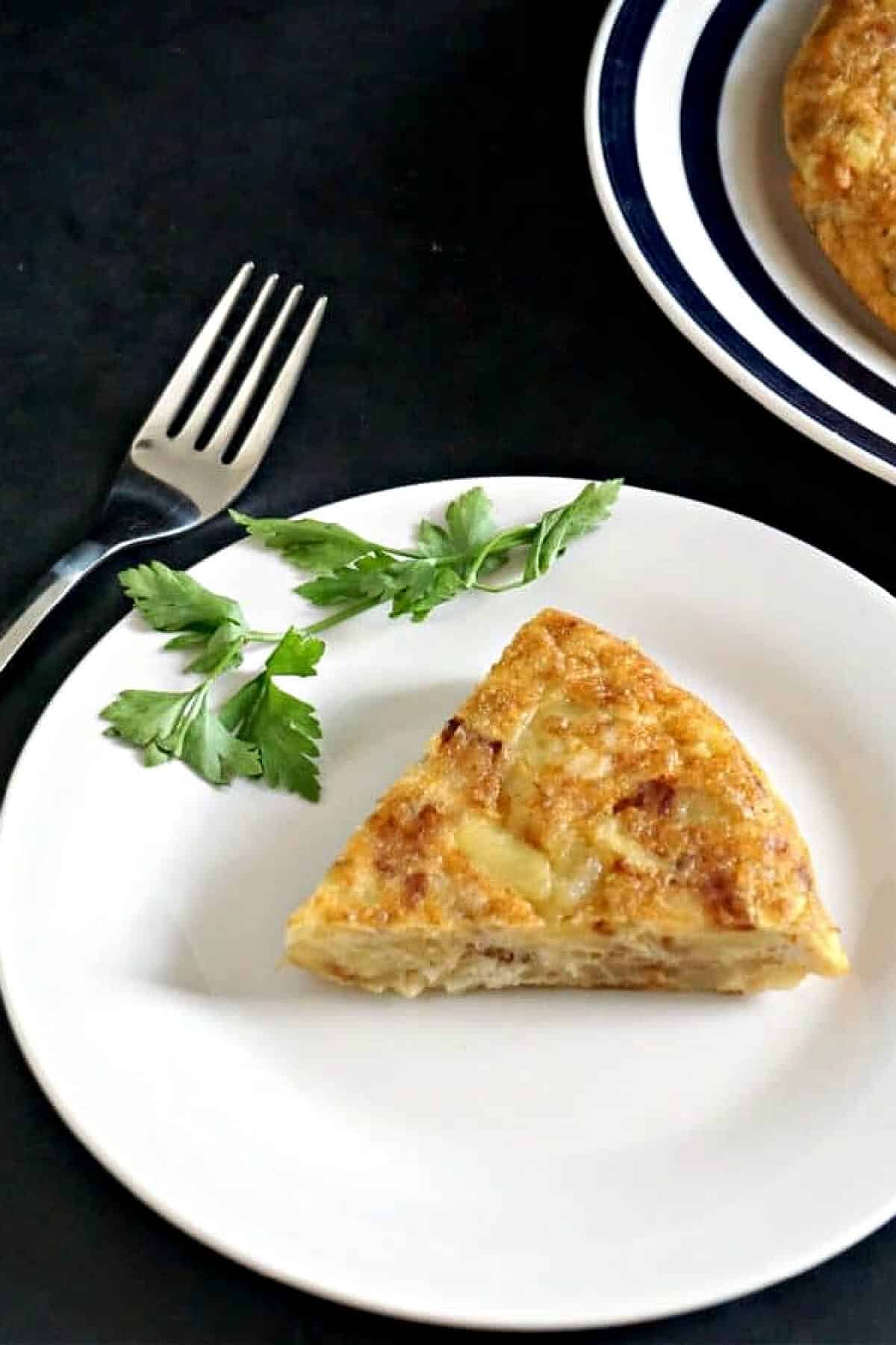 A white plate with a slice of potato omelette and a sprig of parsley on the side