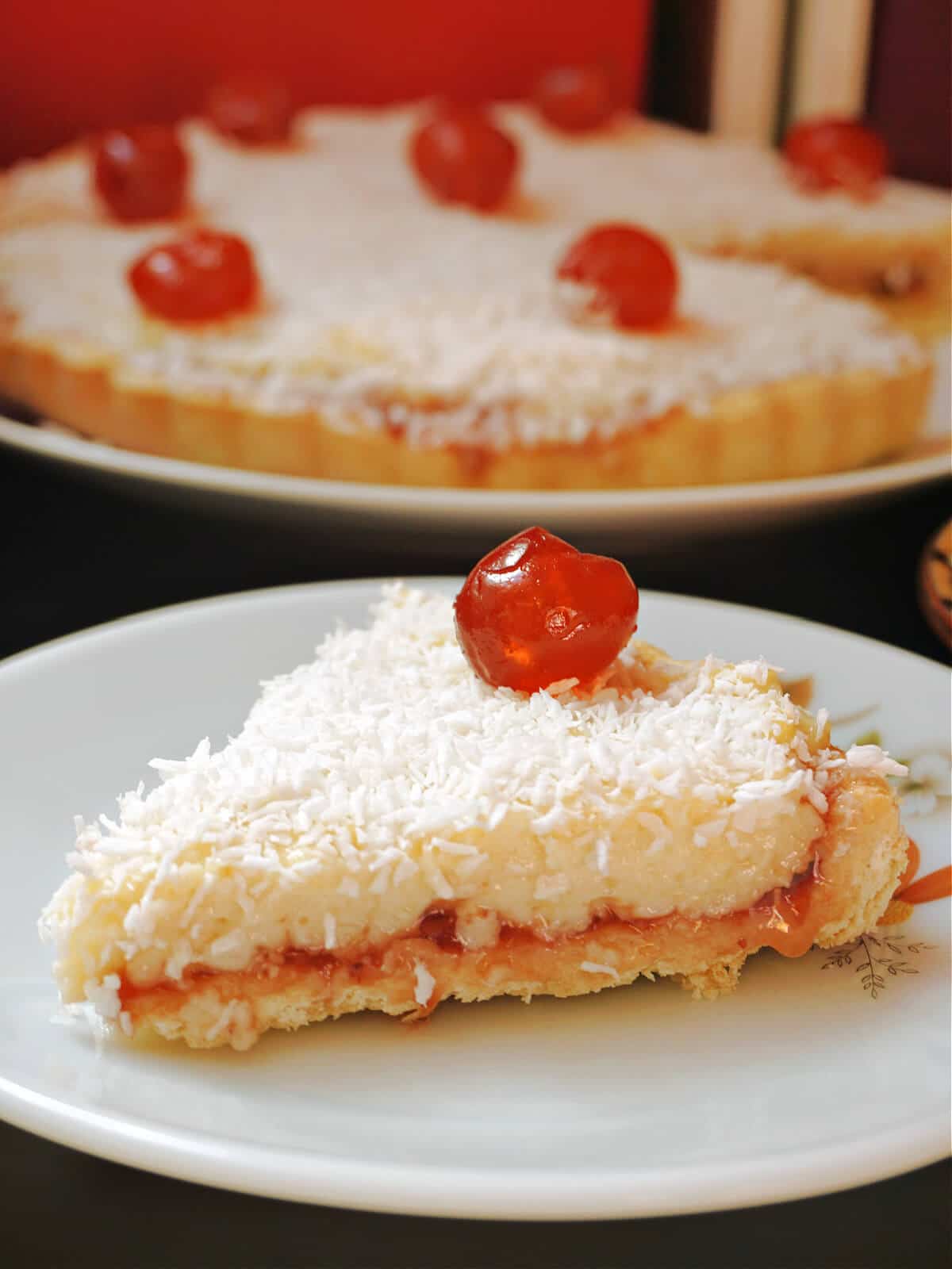 A slice of tart on a white plate with a plate with the rest of the tart in the background.
