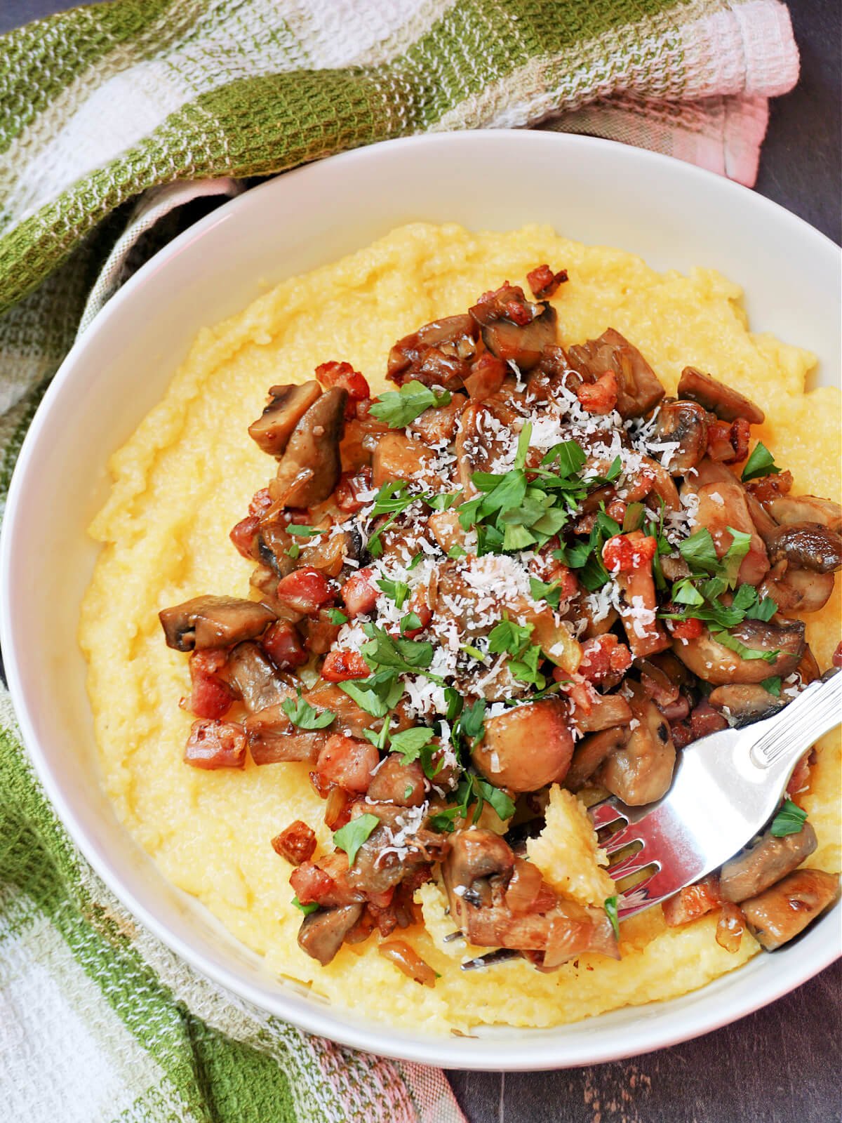 A white bowl with polenta topped with mushrooms, bacon, parmesan and parsley.