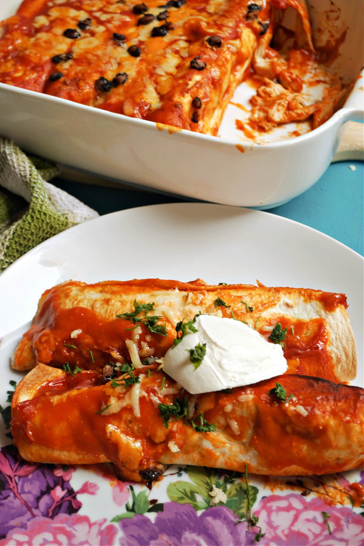 A white plate with 2 enchiladas topped with cream and chopped parsley and a dish with more enchiladas at the top