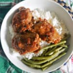 A white bowl with 3 chicken thighs in sauce sitting over a bed of rice and green beans on the side.