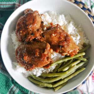A white bowl with 3 chicken thighs in sauce sitting over a bed of rice and green beans on the side.
