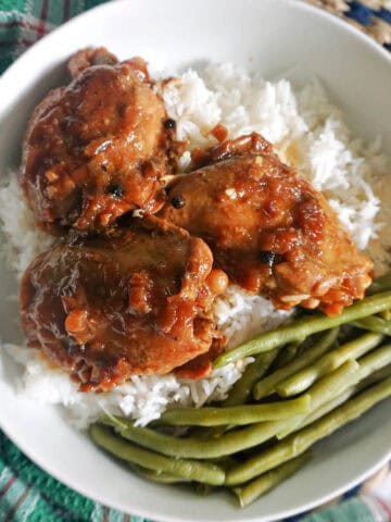 A white bowl with 3 chicken thighs in sauce sitting over a bed of rice and green beans on the side.