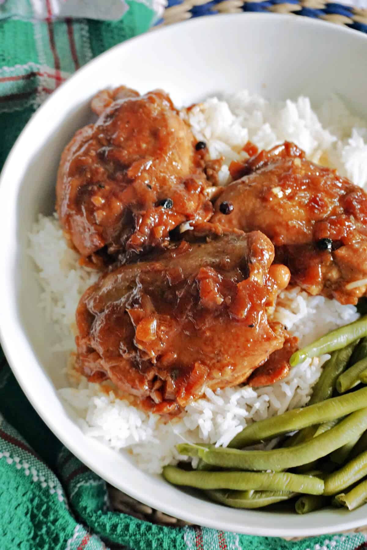 A white bowl with 3 chicken thighs covered in sauce sitting on a bed of rice with green beans on the side.