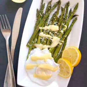 A white rectangle plate with roasted asparagus, poached egg, sauce and 2 slices of lemon