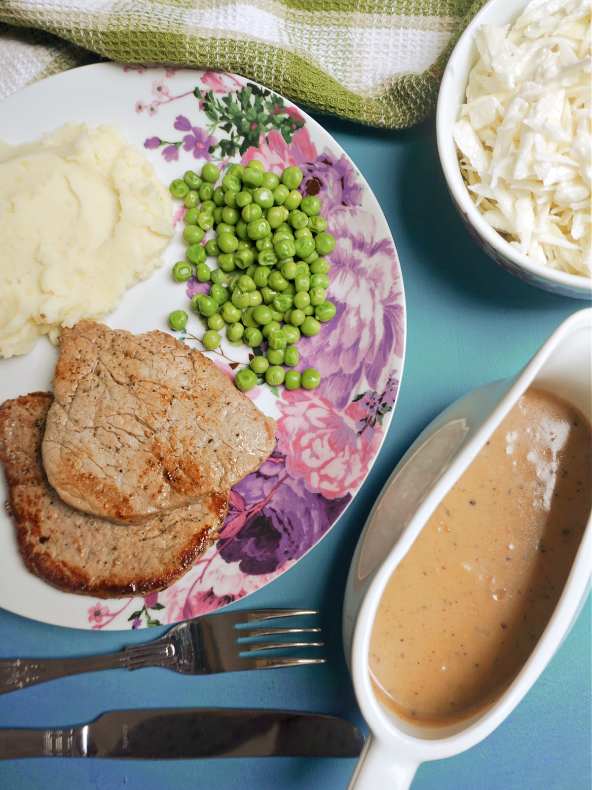 Overhead shoot of a white plate with 2 fillet steaks, mash and peas, and a jug of peppercorn sauce on the side.