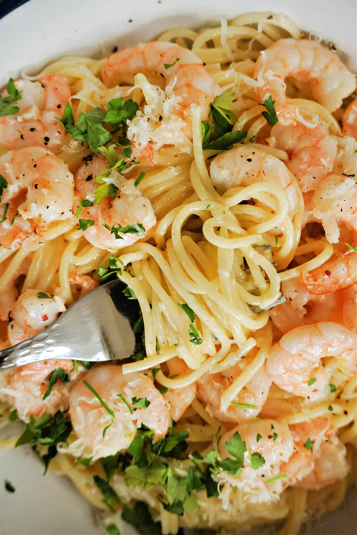 Close-up to a plate with spaghetti and prawns and a fork twisting the spaghettu around. It is garnished with chopped parsley and parmesan.