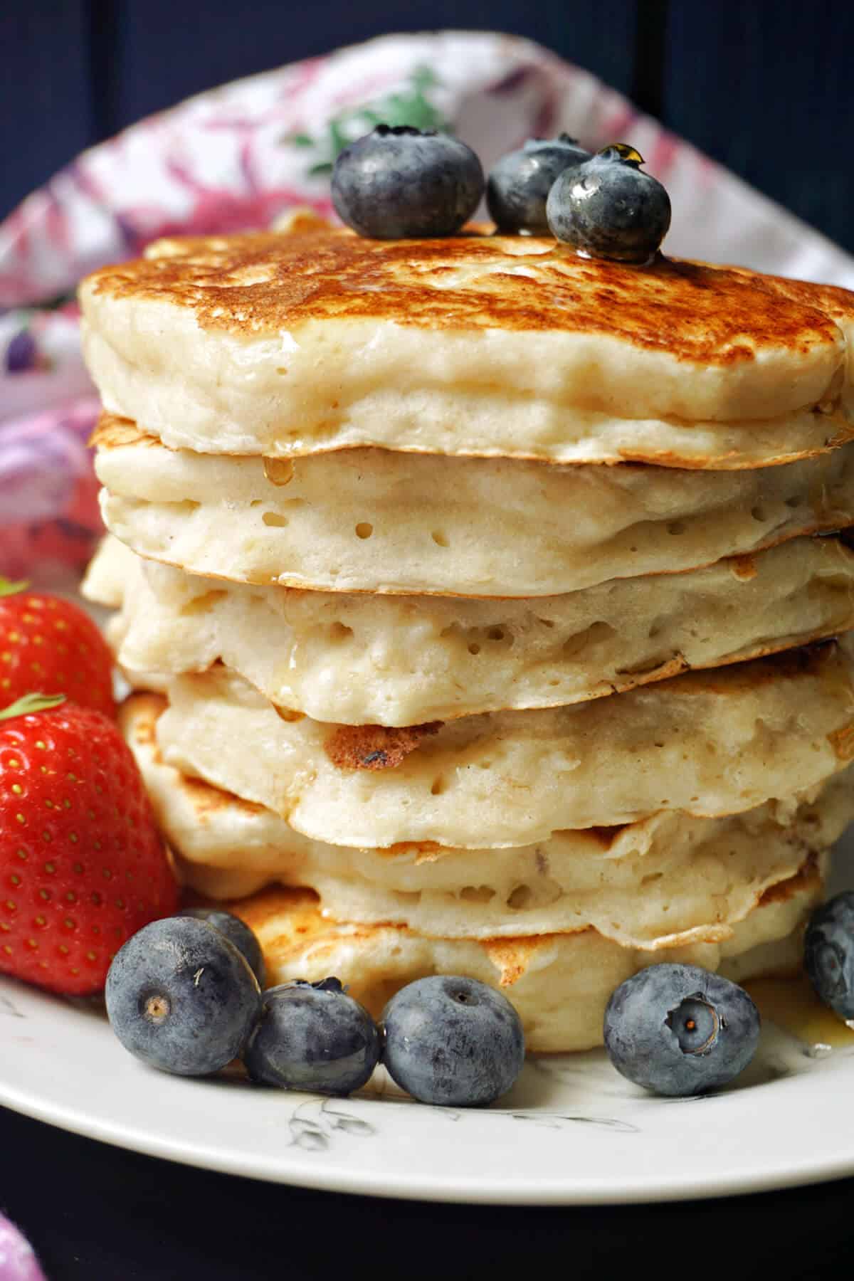 A stack of pancakes with blueberries around and maple syrup pour over them.