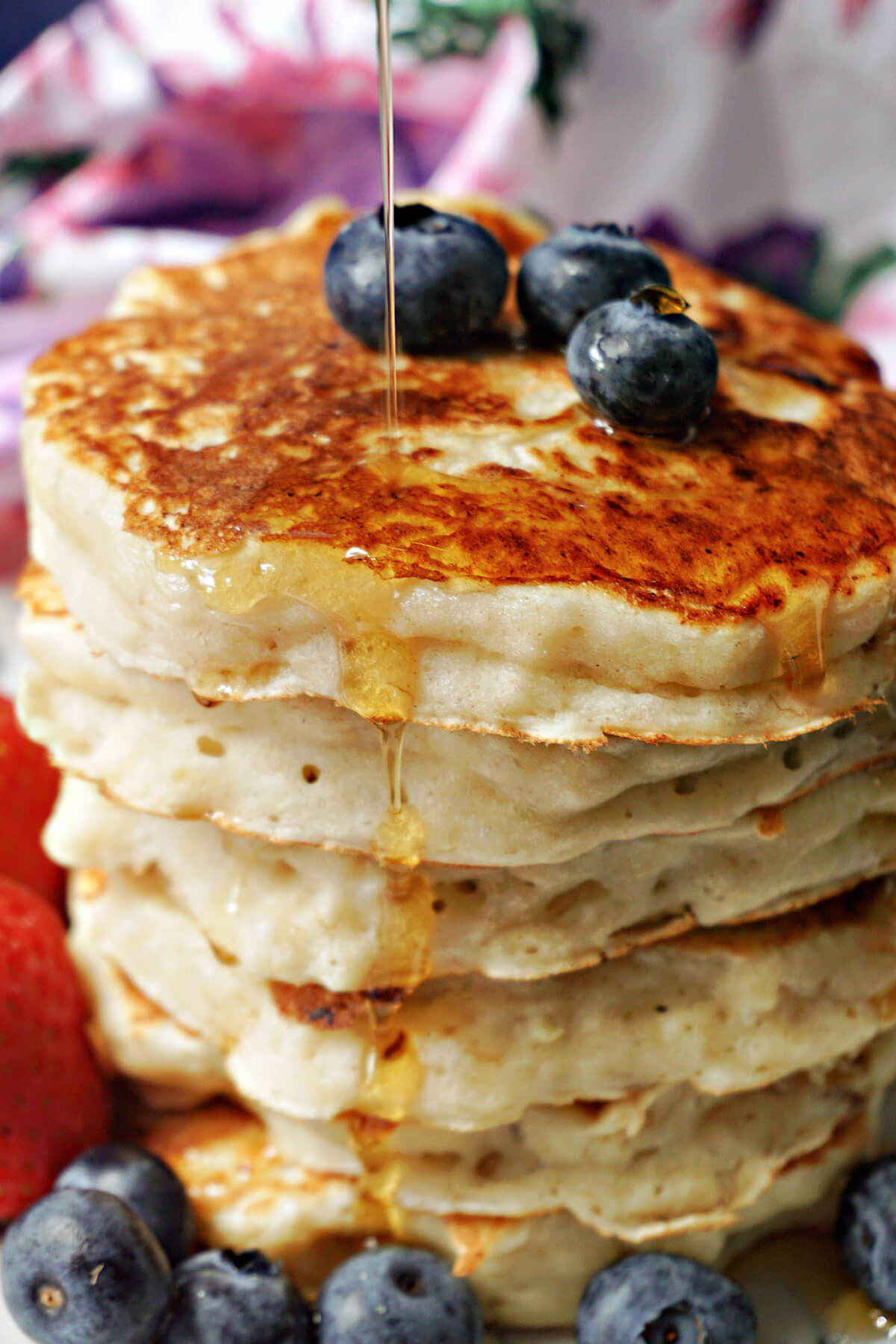 A pile of pancakes being drizzled with maple syrup and topped with blueberries.