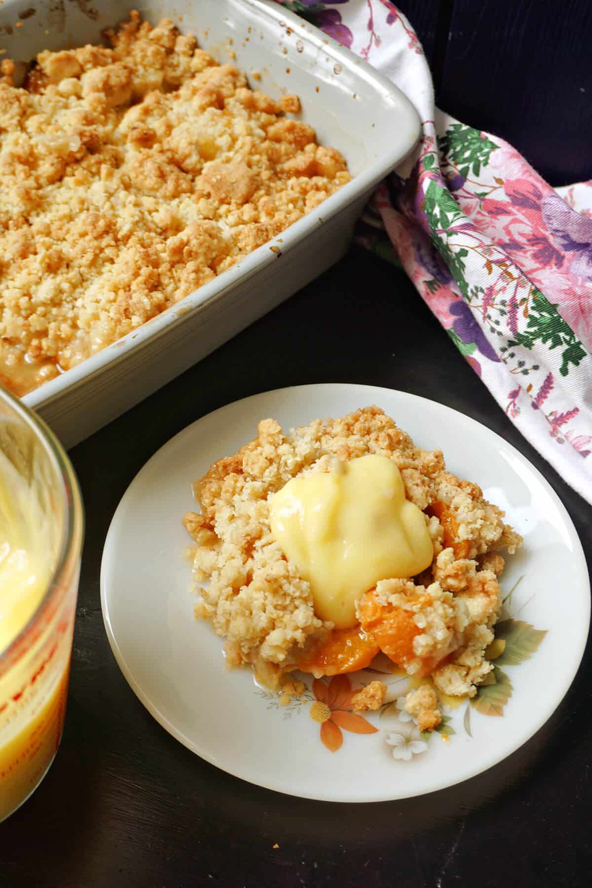 A white plate with a portion of apricot crumble and a dish with more crumble at the top.