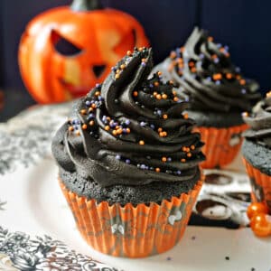 3 black cupcakes on a plate with a spooky pumpkin in the background.