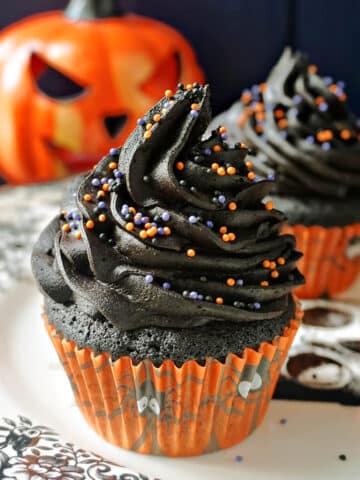 3 black cupcakes on a plate with a spooky pumpkin in the background.