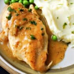 Close-up shoot of a chicken breast smothered in gravy with mash and peas around.