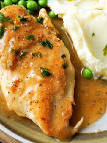 Close-up shoot of a chicken breast smothered in gravy with mash and peas around.