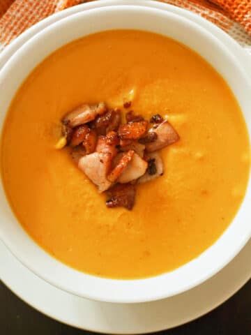 A white bowl with pumpkin and red lentil soup garnished with bacon