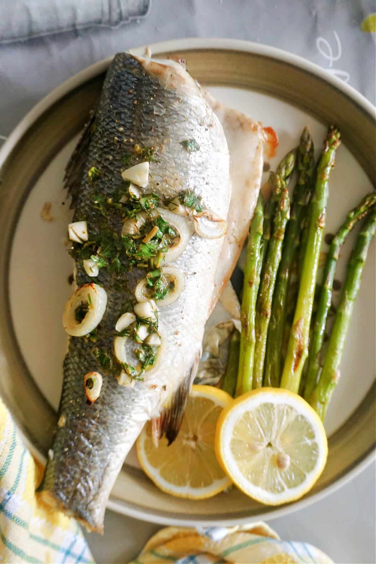 A plate with a baked sea bass topped with lemon butter sauce, asparagus on the side and 2 lemon slices.