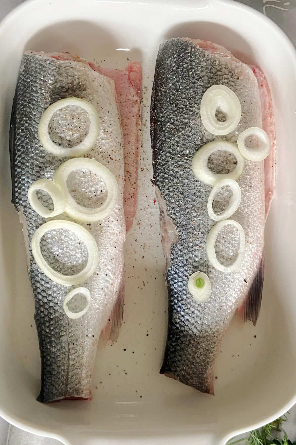 A baking dish with 2 sea bass topped with onion rings.