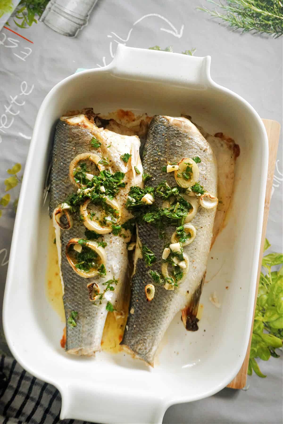 A baking dish with 2 seabass topped with onion rings and herby sauce.