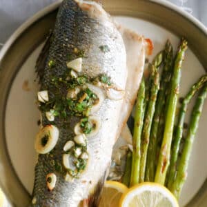 A plate with a whole sea bass, asparagus and 2 lemon slices.