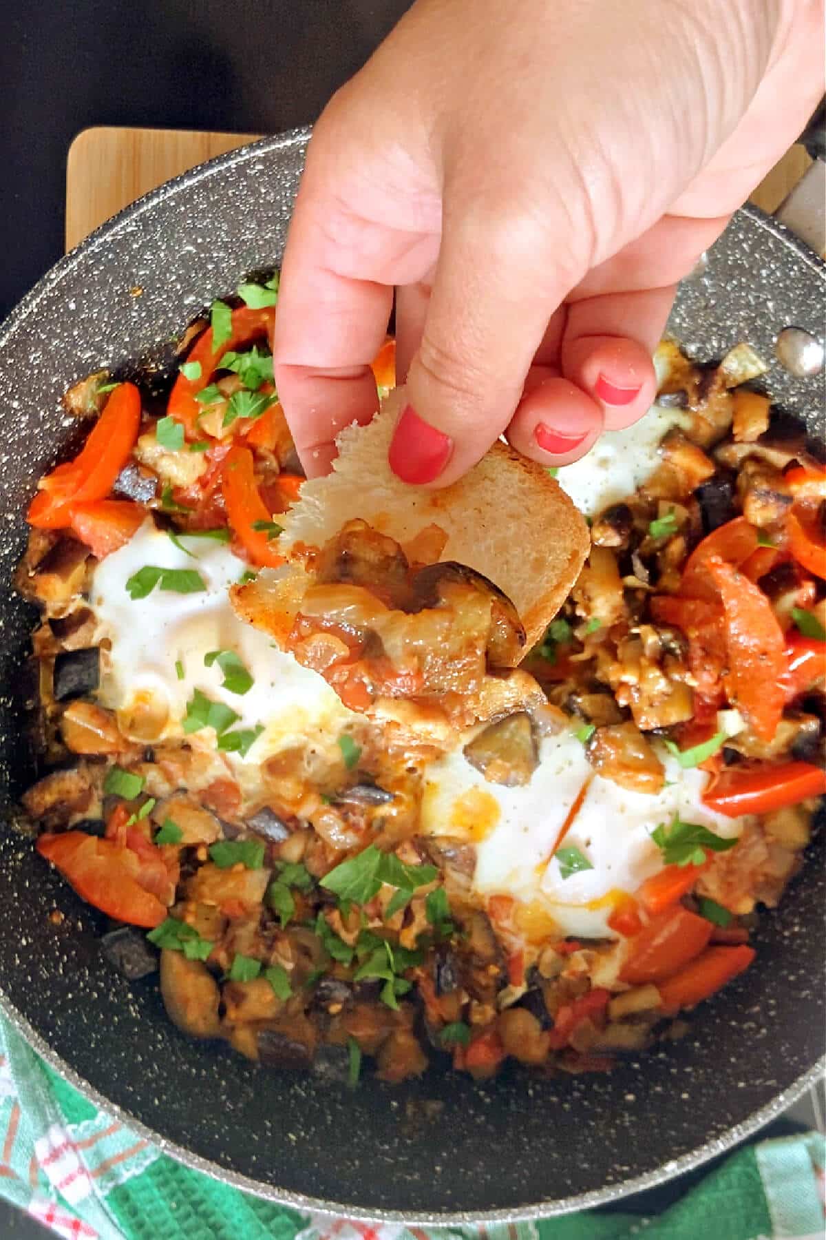 A frying pan with aubergine shakshuka and hand holding a piece of bread with shakshuka on it.