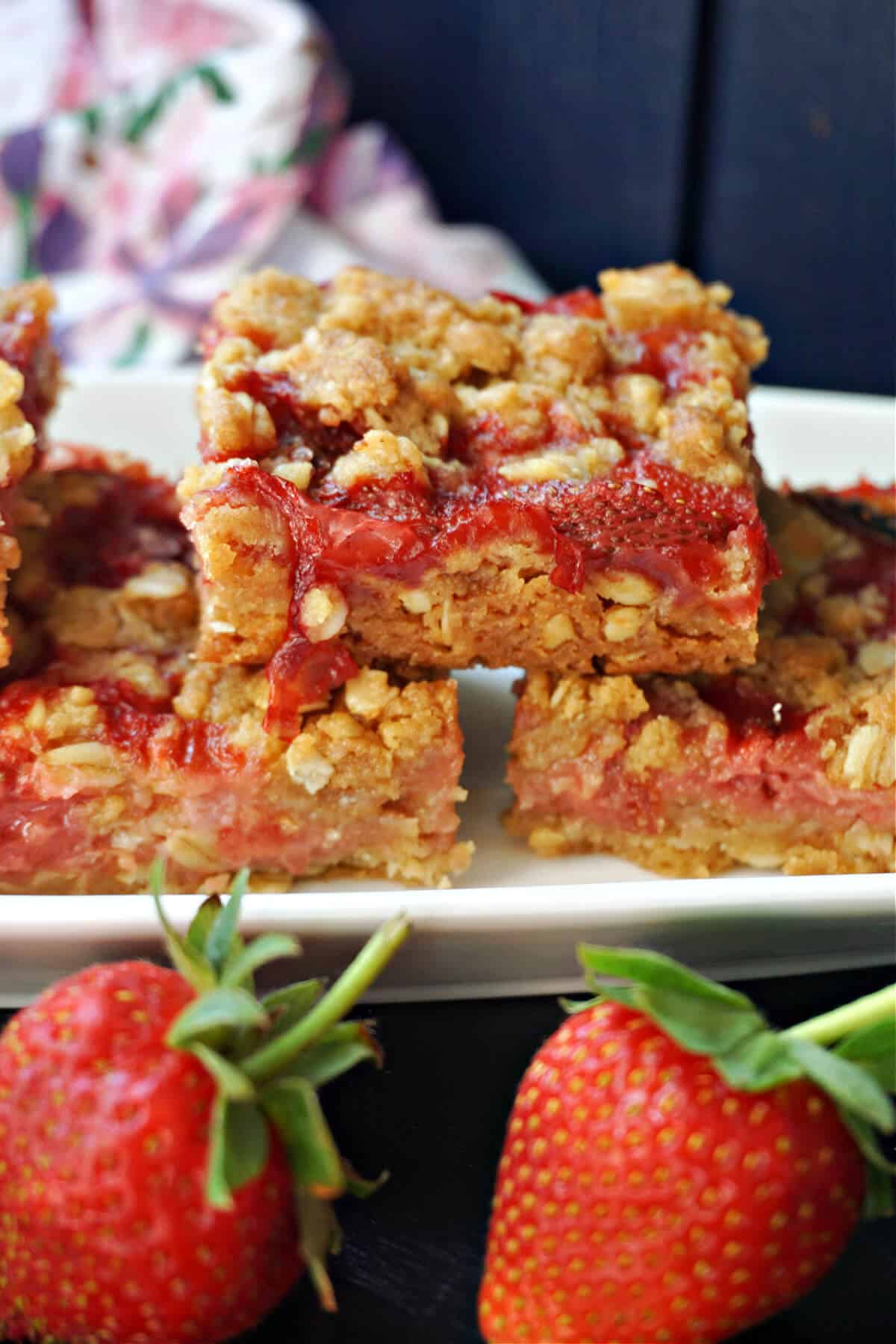 A stack of 3 strawberry crumble bars on a white plate with 2 strawberries in front of it.
