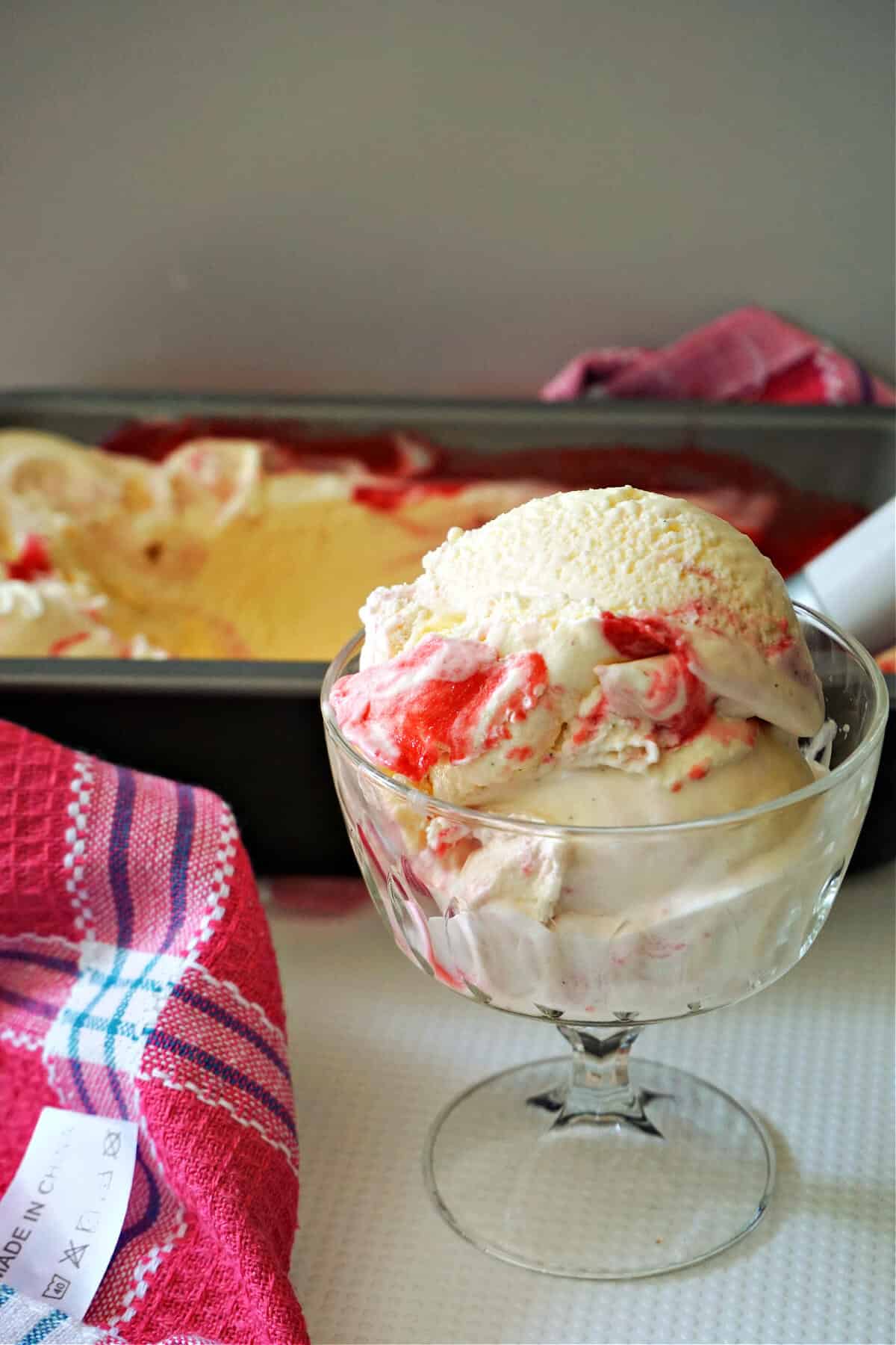 A cup with 2 scoops of raspberry ripple ice cream with a dish with more ice cream in the background.
