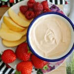 A white plate with a small bowl of peanut butter fruit dip, strawberries, slices of apples and raspberries around it.