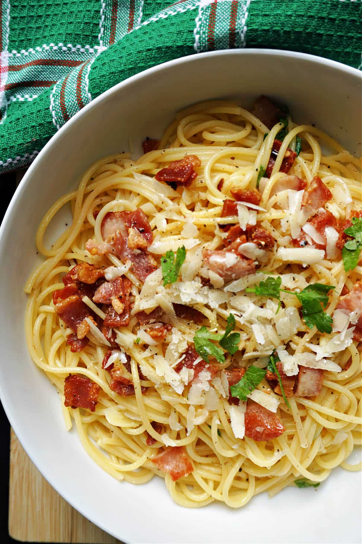 A white bowl with spaghetti topped with bacon, parmesan and chopped parsley.
