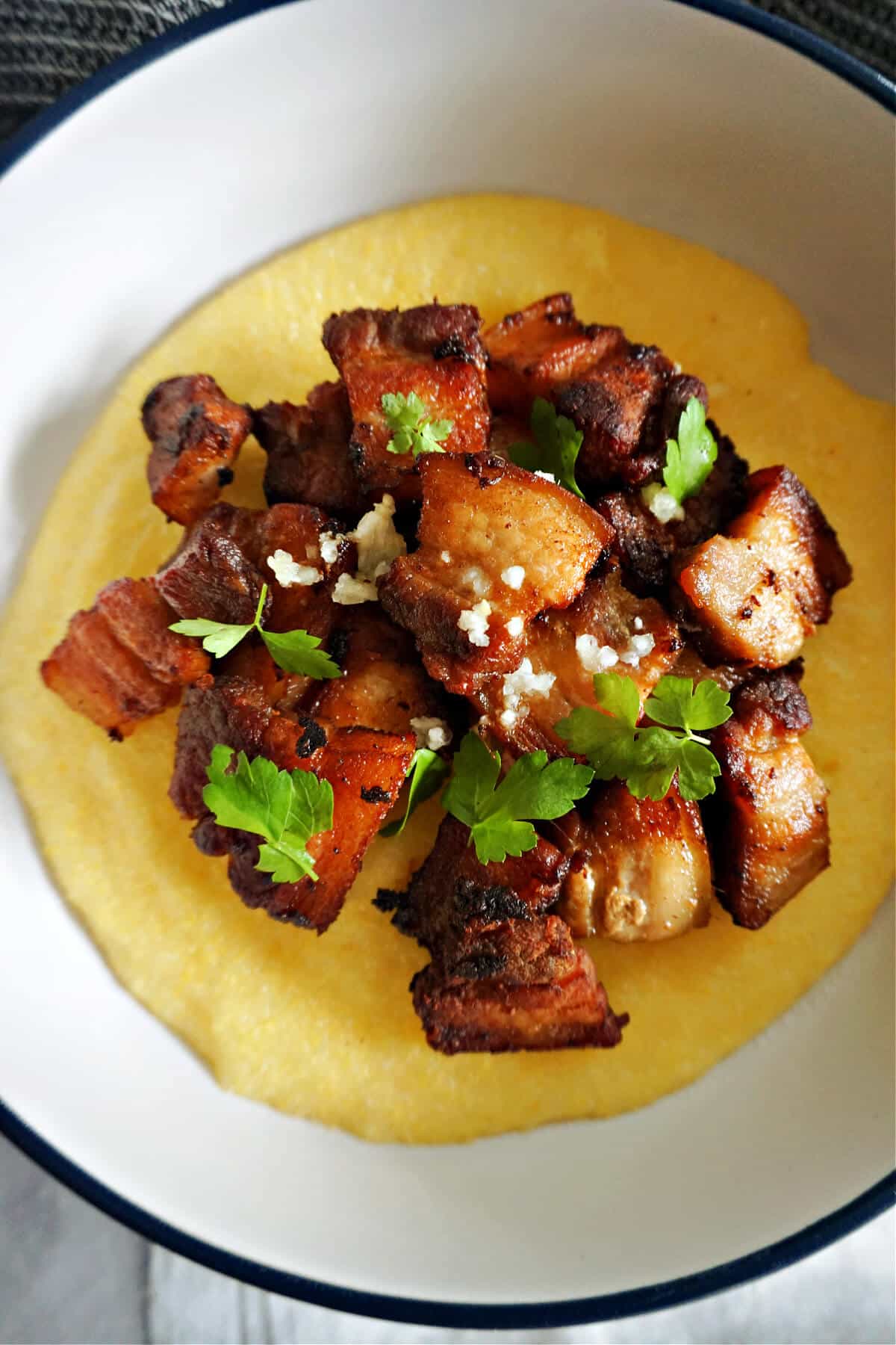 A white plate with polenta topped with pork belly bites garnished with garlic and parsley.