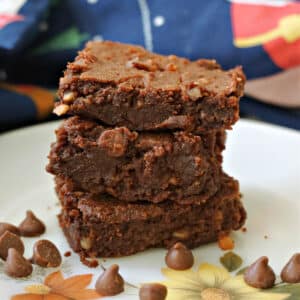 A stack of 3 brownies on a plate with chocolate chips around them.