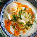 A blue plate with 2 poached eggs on a bed of yogurt with spicy sauce and fresh herbs on top.