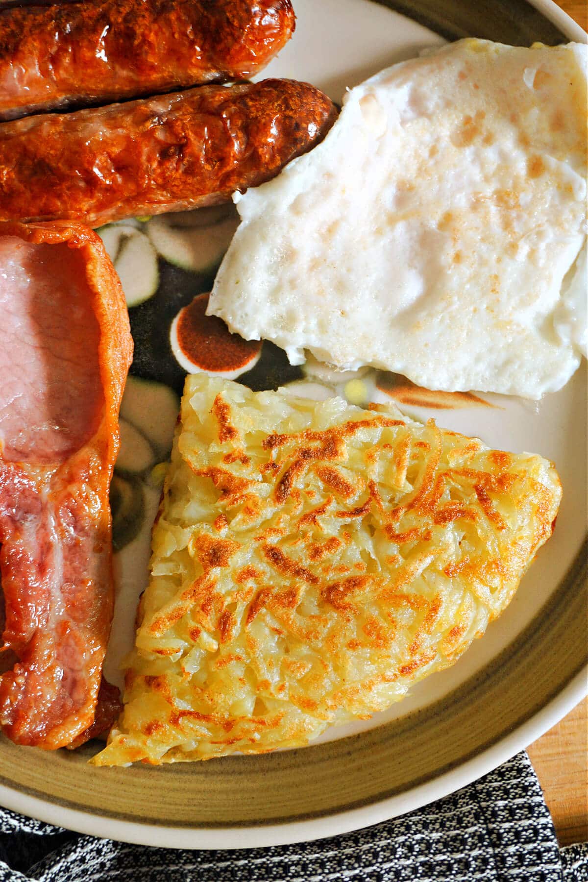 A slice of rosti, a fried egg, 2 sausages and bacon on a plate.