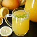 A glass of mango lemonade with 2 slices of lemon and a mango around it.