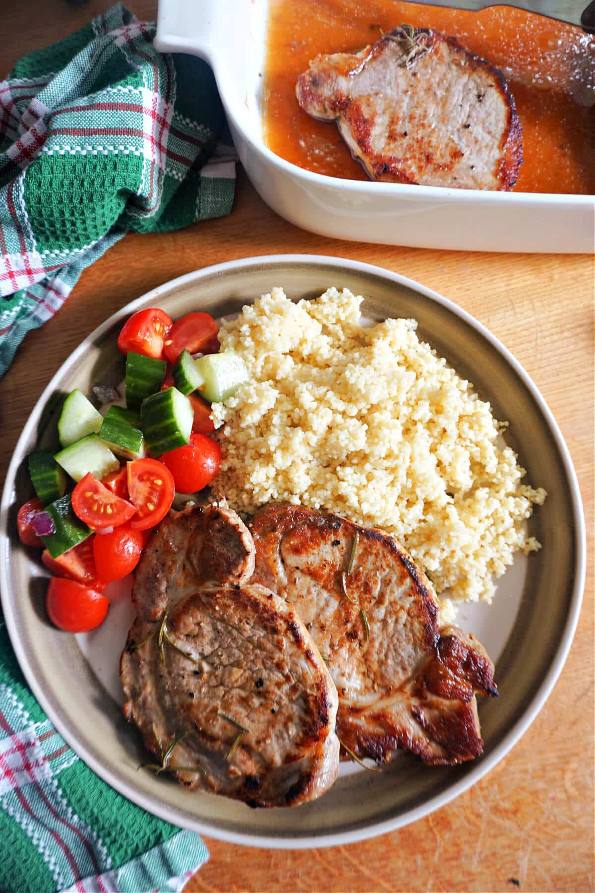 A plate with 2 pork steaks, couscous and a cucumber and tomato salad and a dish at the top with more steaks.