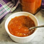 Peach jam in a white ramekin with a teaspoon in it and a jar of jam at the back.