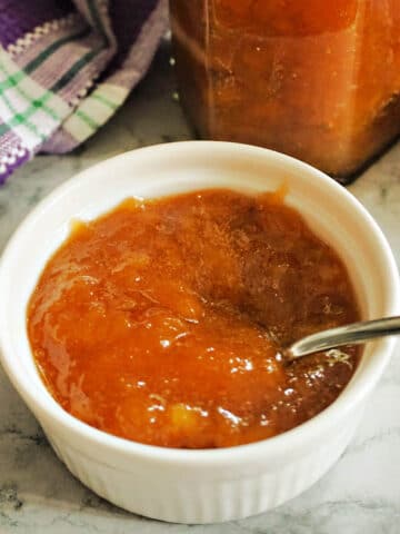 Peach jam in a white ramekin with a teaspoon in it and a jar of jam at the back.