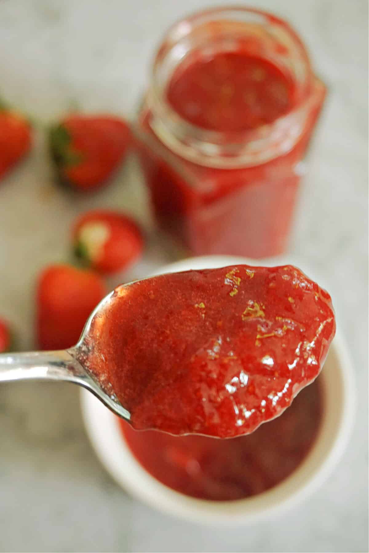 A spoonful of strawberry jam over a ramekin of more jam and a jar and fresh strawberries in the background.