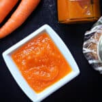 A white ramekin with carrot jam, 2 carrots and a jar of jam on the table.