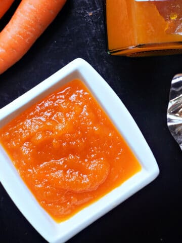 A white ramekin with carrot jam, 2 carrots and a jar of jam on the table.