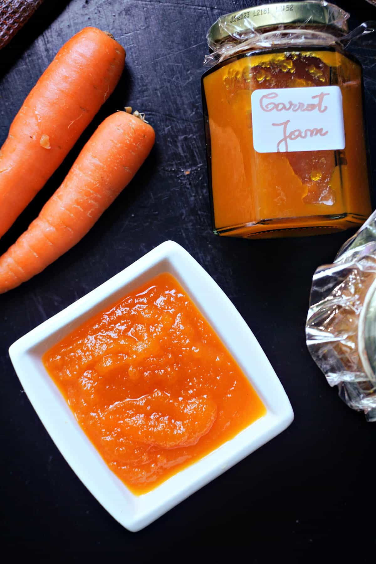 A white ramekin with carrot jam, 3 carrots and a carrot jam jar on a black table.