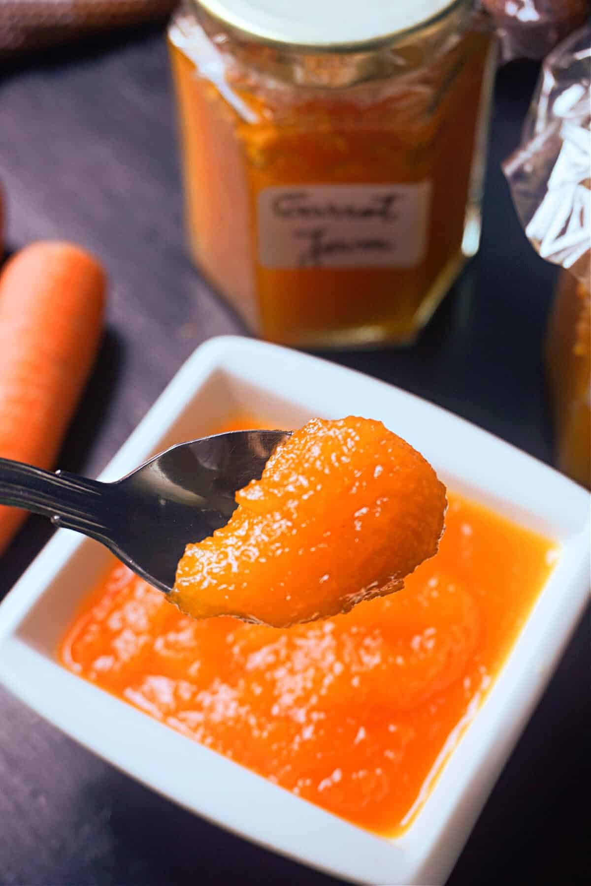 A spoonful of carrot jam over a white ramekin with more jam, a carrot and a jam jar on the table.
