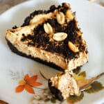 A slice of peanut butter pie with oreo crust on a plate with a dessert spoon of pie on it.