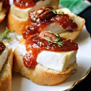A white plate with 2 crostini topped with bre, fig jam, pecans and thyme, with other crostini partially visible.