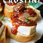 A plate with 2 crostini topped with brie and fig jam, pecans and thyme with other crostini partially visible.