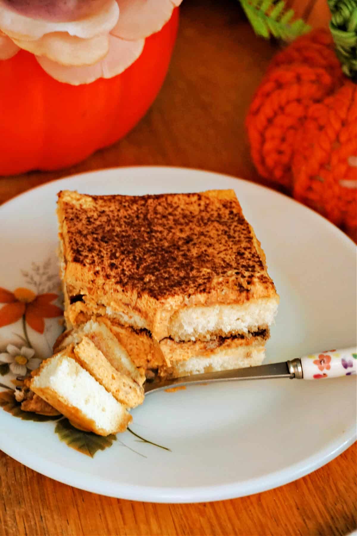 A slice of pumpkin tiramisu on a white flowery plate with a bite of tiramisu on a fork.