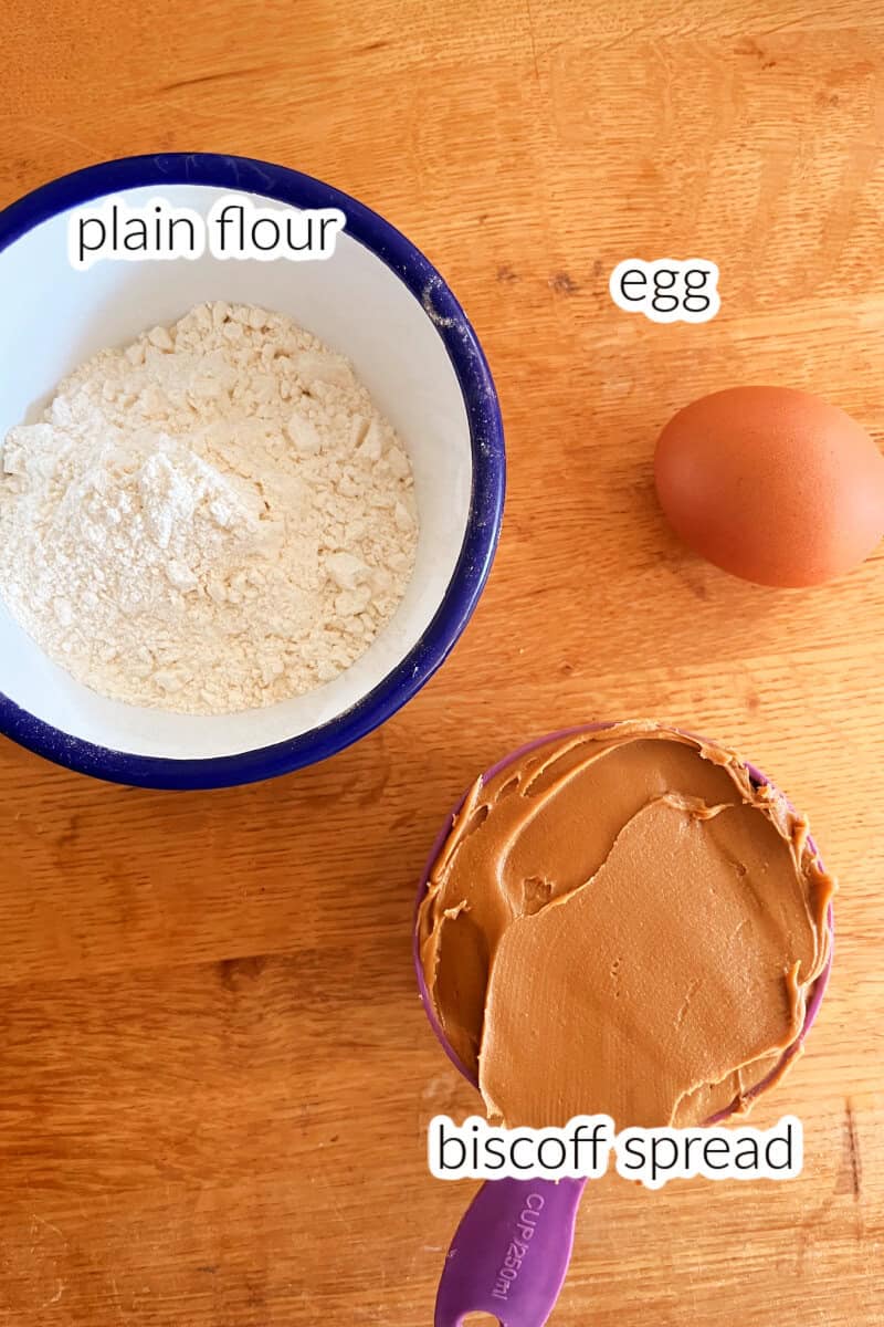 A white and blue ramekin with flour, an egg and a cup of peanut butter on a wooden table.